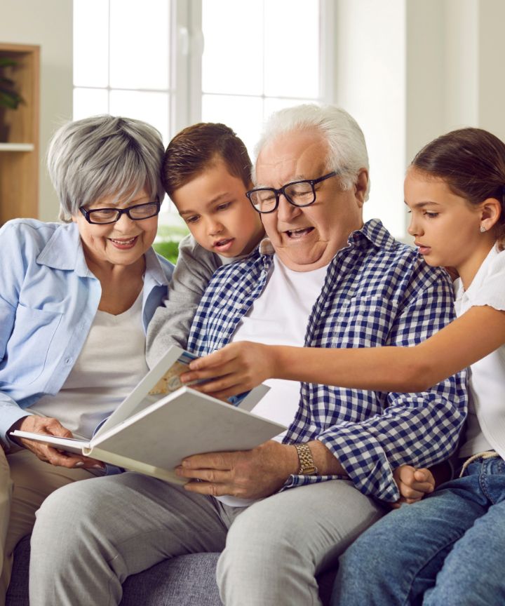 Grandfather reading a book to his two young grandchildren, joined by their grandmother.